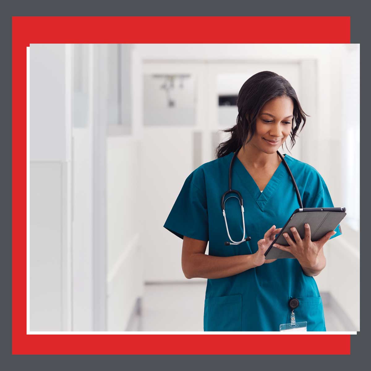 Smiling nurse in teal scrubs reviewing patient information on a tablet in a brightly lit medical hallway
